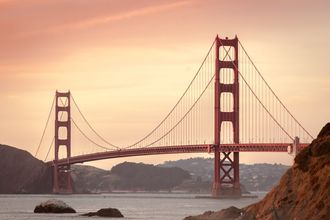 The Golden Gate Bridge seen from the rocky shoreline at sunset, both towers spanning the bay